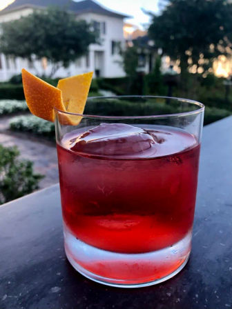 Red cocktail in a rocks glass with large ice sphere and orange peel garnish on an outdoor patio table at dusk, garden and house blurred in background