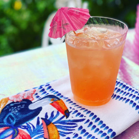 Iced orange tropical cocktail in a plastic cup with a pink paper umbrella, set on a colorful toucan beach towel for a bright summer patio vibe