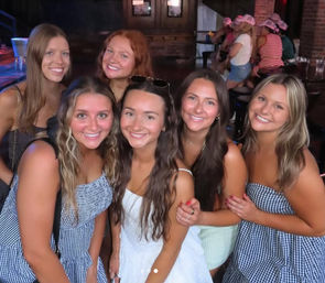 Six friends smiling and posing inside a dimly lit bar, wearing summer dresses with one in white at center — group night out photo.
