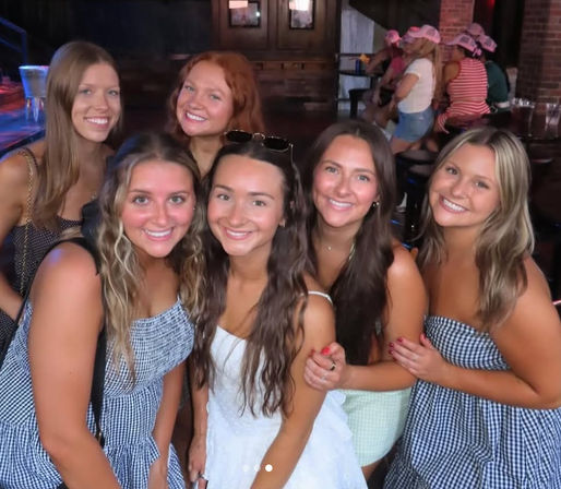 Six friends smiling and posing inside a dimly lit bar, wearing summer dresses with one in white at center — group night out photo.