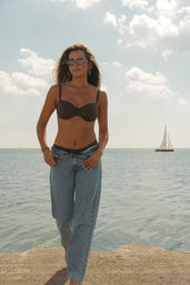 Woman in a black bikini top and loose denim jeans walking on a concrete pier by the sea, wearing sunglasses and hoop earrings with a sailboat on the horizon under a sunny sky.