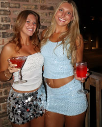 Two smiling friends in sparkly summer outfits holding red cocktails on a nighttime outdoor patio with a brick wall backdrop