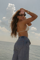 Woman in white crochet bikini top and high-waist jeans wearing sunglasses and gold jewelry posing beachside by the ocean under a sunny sky with puffy clouds