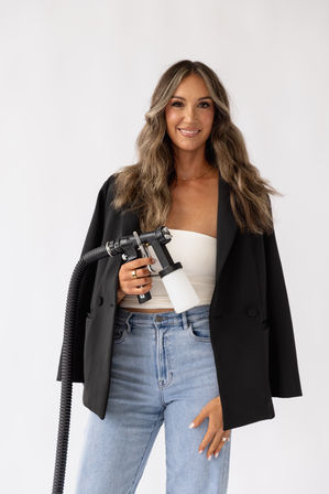 Smiling woman with long wavy hair wearing a black blazer and light jeans holding a handheld spray gun with hose and white solution bottle in a clean studio portrait