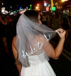 Woman in white dress holding a veil embroidered "bride to be" on a neon-lit downtown street at night — lively bachelorette party vibe.