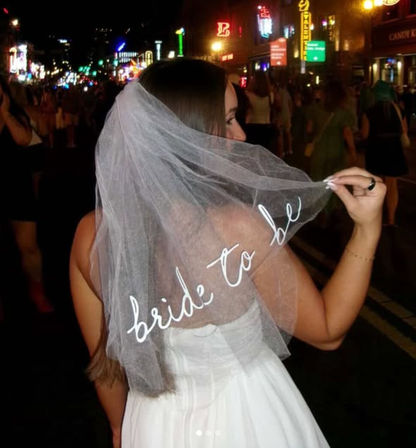 Woman in white dress holding a veil embroidered "bride to be" on a neon-lit downtown street at night — lively bachelorette party vibe.