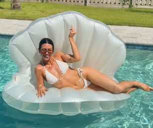 Person in white bikini lounging on inflatable shell float in a sunlit backyard pool, smiling and playfully flipping the middle finger.