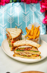 Grilled club sandwich stacked with turkey, bacon, lettuce, tomato and pickles, skewered and served with golden fries in a small metal basket on a white plate against a turquoise patterned patio cushion with pink flowers.