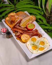 Hearty tropical breakfast platter on a white rectangular plate with four sunny-side-up eggs, crispy bacon and sliced sausages, buttered toast and French toast, small bowls of guacamole and spread, and jam jars, photographed against green palm leaves.