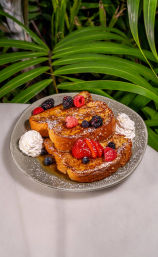 Golden French toast stack dusted with powdered sugar, drizzled with syrup and topped with strawberries, raspberries, blackberries and blueberries, served with whipped cream on a ceramic plate against lush tropical leaves — vibrant brunch scene.