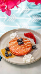 Bright red pancakes dusted with powdered sugar, stacked on a beige plate and topped with blueberries and blackberries, with syrup, whipped cream and sliced strawberries on a turquoise patterned tablecloth — colorful brunch dish.