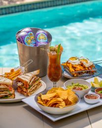 Poolside brunch spread: stacked club sandwiches and fries, tortilla chips with guacamole and salsa, loaded fries topped with sunny-side eggs, a Bloody Mary-style cocktail garnished with olives and celery, and a bucket of canned craft beer by a turquoise swimming pool.