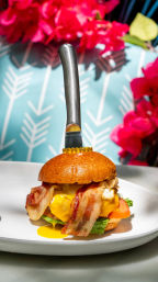 Crispy bacon and melted cheddar breakfast sandwich with runny egg on a toasted brioche bun, served on a white plate with a knife inserted through the bun and bright floral backdrop — brunch dish.