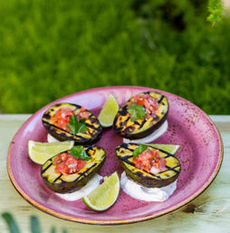 Grilled avocado halves with char marks, filled with pico de gallo and cilantro, on dollops of crema with lime wedges on a pink plate outdoors