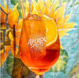 Bright orange spritz cocktail with ice, a fresh orange slice and straw in a stemmed glass against a colorful sunflower tile mural backdrop — refreshing summer aperitif vibe