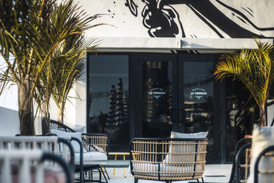 Sunlit rooftop terrace with rattan lounge chairs, palm trees and modern outdoor seating facing glass doors reflecting a city skyline