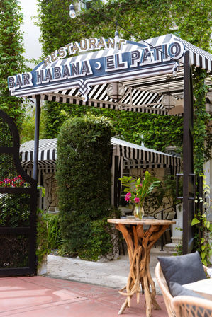 Garden restaurant patio entrance with black-and-white striped awning and marquee sign, lush green vines, string lights, and a rustic driftwood high table topped with a vase of pink flowers beside wicker seating.