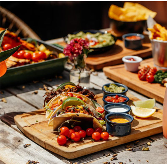 Vibrant street-style tacos with lime wedges, cherry tomatoes and three dipping sauces served on a wooden board on an outdoor patio table, chips and fries in the blurred background.