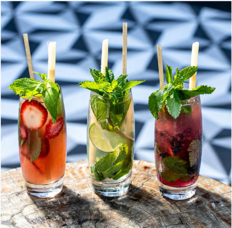Three colorful summer cocktails on a weathered wooden table — strawberry-pink drink with sliced berries and mint, a clear lime-and-mint cooler with lime wheels, and a purple blackberry-mint mocktail, each garnished with fresh mint and bamboo straws against a geometric tiled patio backdrop.