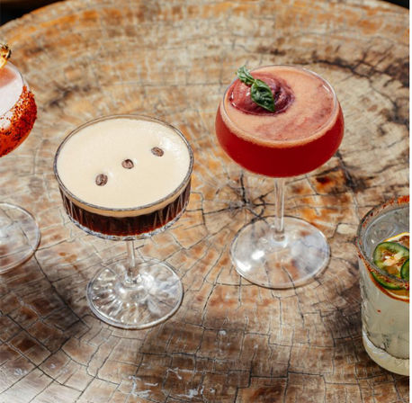 Colorful craft cocktails on a rustic round wood table: an espresso martini with three coffee beans, a bright red fruit coupe garnished with a basil leaf, a chili‑rimmed cocktail, and a jalapeño‑topped clear drink.