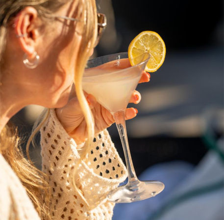 Person with blonde hair in a knit sweater sipping a lemon‑garnished martini on a sunlit outdoor patio — a refreshing, summery moment.