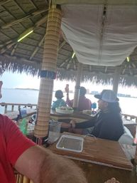 Waterfront tiki bar interior with thatched roof and wrapped wooden posts, people seated at a wooden bar with snacks and drinks, one guest holding up a phone toward the bay and distant island skyline.
