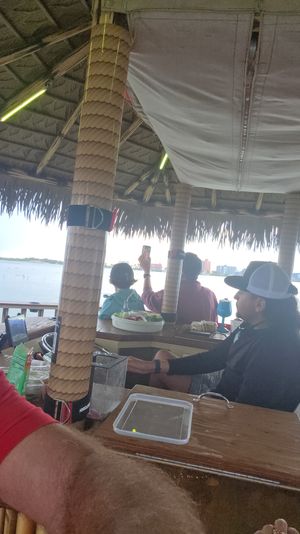 Waterfront tiki bar interior with thatched roof and wrapped wooden posts, people seated at a wooden bar with snacks and drinks, one guest holding up a phone toward the bay and distant island skyline.