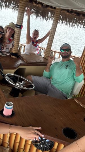 Group of friends in red-white-blue leis laughing and taking a selfie aboard a thatched-roof tiki bar boat on calm water, seated around a wooden bar with drinks and a man at the helm flashing a shaka sign.