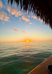 Tropical sunset over calm turquoise ocean, golden sun reflecting on rippled water beneath a palm-thatched roof with a wooden deck edge and a small sailboat on the distant horizon.