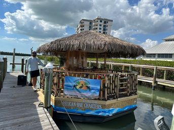 Tiki-roofed charter boat moored at a bayfront wooden dock in a tropical marina, a person in sunglasses stands on the dock with low-rise condos, mangroves and puffy clouds under a blue sky in the background.