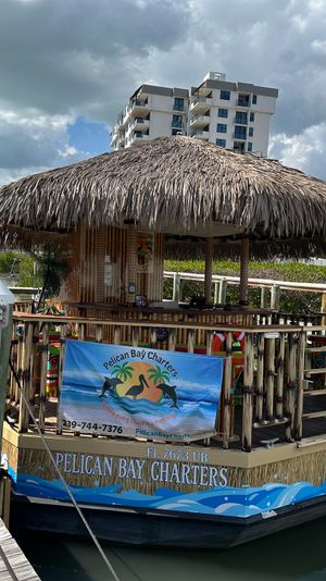 Tiki-roofed charter boat moored at a bayfront wooden dock in a tropical marina, a person in sunglasses stands on the dock with low-rise condos, mangroves and puffy clouds under a blue sky in the background.