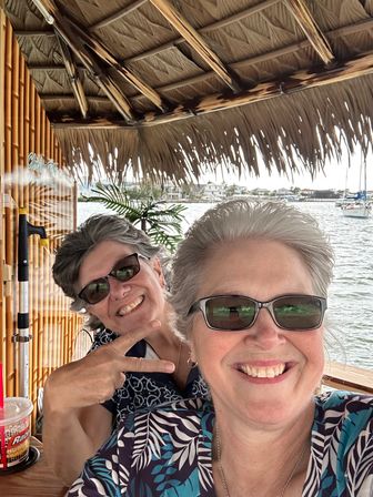Two smiling women in sunglasses take a cheerful selfie under a thatched tiki hut at a waterfront marina with boats and palms in the background.