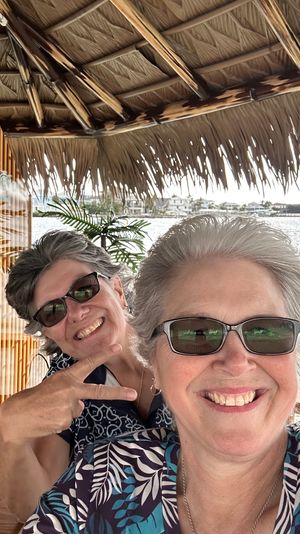 Two smiling women in sunglasses take a cheerful selfie under a thatched tiki hut at a waterfront marina with boats and palms in the background.