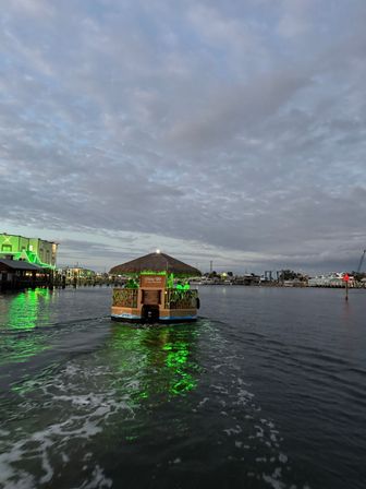 Tiki-style party boat with a thatched roof cruising through a marina at dusk, green lights reflecting on calm harbor water beneath a cloudy sky.