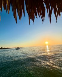 Tropical sunset over calm ocean with golden sun reflecting on rippling water, a small motorboat near the distant shoreline and a thatched-roof overhang framing the view.