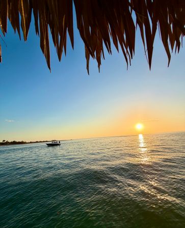Tropical sunset over calm ocean with golden sun reflecting on rippling water, a small motorboat near the distant shoreline and a thatched-roof overhang framing the view.