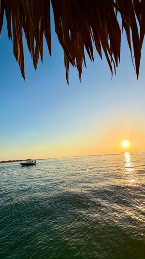 Tropical sunset over calm ocean with golden sun reflecting on rippling water, a small motorboat near the distant shoreline and a thatched-roof overhang framing the view.