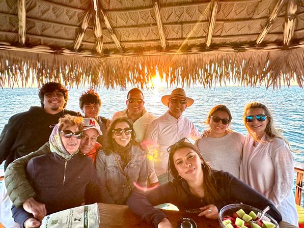 Smiling multi‑generation group gathered under a thatched tiki hut at an oceanfront sunset, sunglasses and sun hat, bowl of fresh melon on the table — beachy, coastal scene.