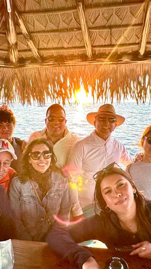 Smiling multi‑generation group gathered under a thatched tiki hut at an oceanfront sunset, sunglasses and sun hat, bowl of fresh melon on the table — beachy, coastal scene.