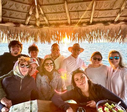 Smiling multi‑generation group gathered under a thatched tiki hut at an oceanfront sunset, sunglasses and sun hat, bowl of fresh melon on the table — beachy, coastal scene.