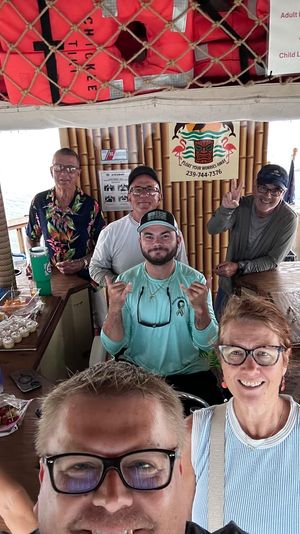 Cheerful group of adults on a bamboo-roof tiki boat over calm water, life vests stored overhead and snacks and drinks on a wooden bar — casual waterfront boat party.