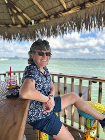Smiling woman in sunglasses relaxing at a waterfront tiki-hut bar, seated on a colorful tiki stool with a drink on the wooden counter and a calm turquoise bay under a blue sky with clouds in the background.