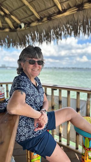 Smiling woman in sunglasses relaxing at a waterfront tiki-hut bar, seated on a colorful tiki stool with a drink on the wooden counter and a calm turquoise bay under a blue sky with clouds in the background.