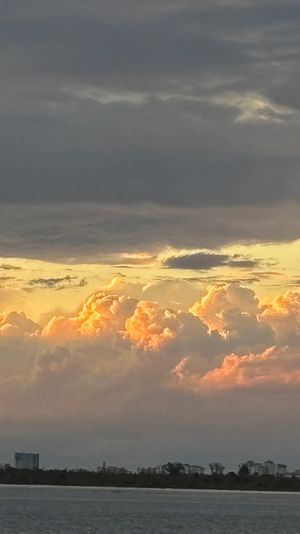 Coastal sunset over calm water with golden-orange clouds glowing beneath dark storm clouds and a silhouetted shoreline