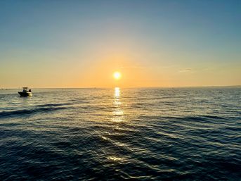 Golden-hour ocean sunset with the sun low on the horizon, shimmering reflection across calm rippled water and a small motorboat silhouetted to the left.