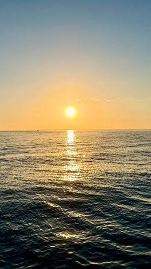 Golden-hour ocean sunset with the sun low on the horizon, shimmering reflection across calm rippled water and a small motorboat silhouetted to the left.