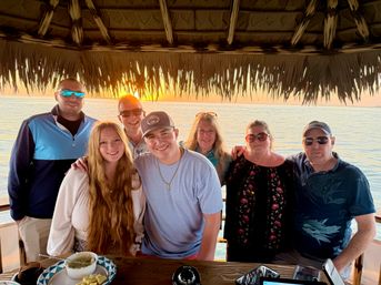 Smiling multi-generational group posing under a thatched tiki hut at sunset over a calm ocean, wooden table with snacks in the foreground.
