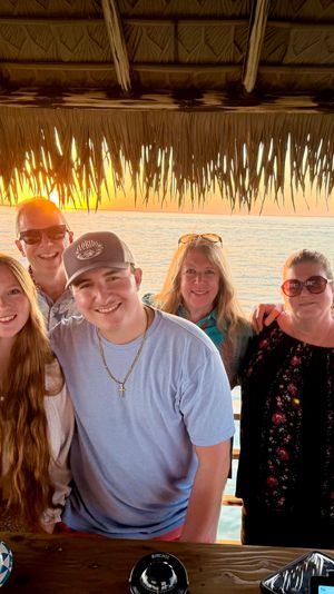 Smiling multi-generational group posing under a thatched tiki hut at sunset over a calm ocean, wooden table with snacks in the foreground.