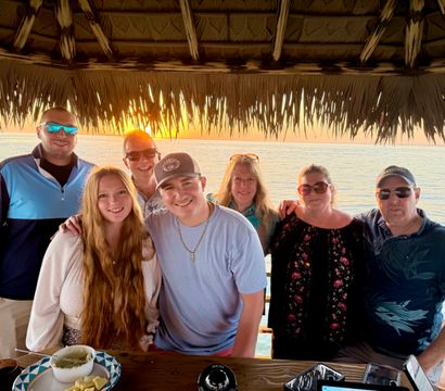 Smiling multi-generational group posing under a thatched tiki hut at sunset over a calm ocean, wooden table with snacks in the foreground.