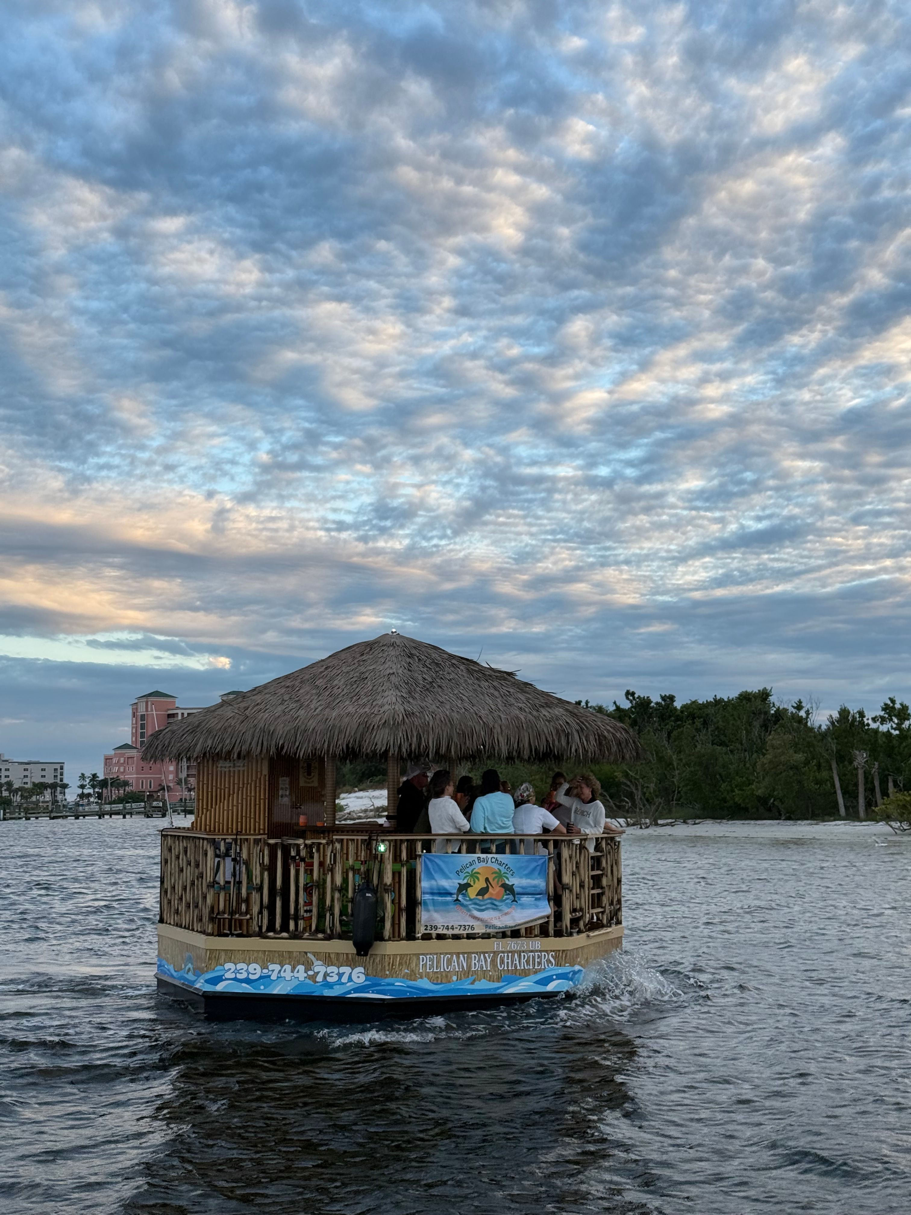 Thatched-roof tiki boat with passengers cruising a coastal inlet near a palm-lined shore under a dramatic, cloud-filled sunset sky.
